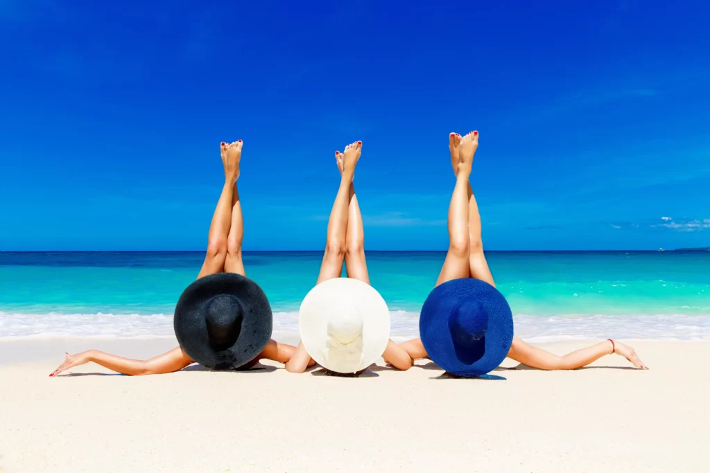 Three young women in straw hats lying on a tropical beach, stretching up slender legs. Blue sea in the background. Summer vacation concept.